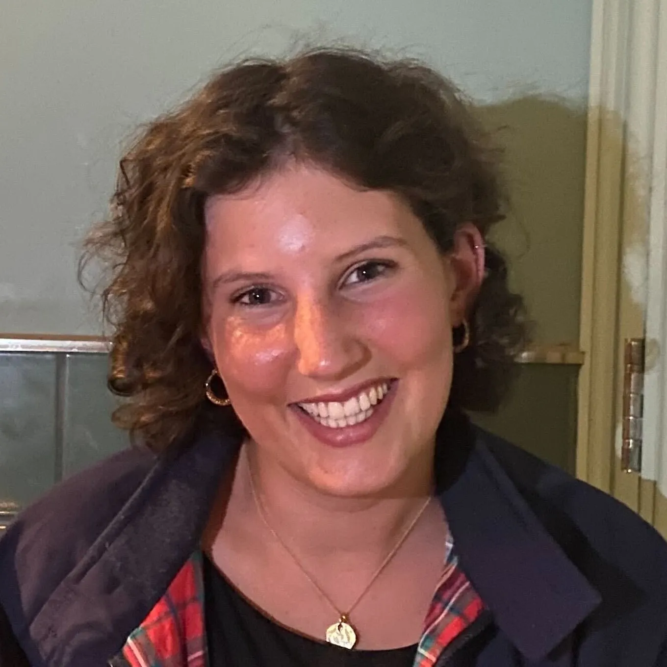 Smiling woman with curly hair wearing gold hoop earrings and a necklace, dressed in a dark jacket with red plaid lining.