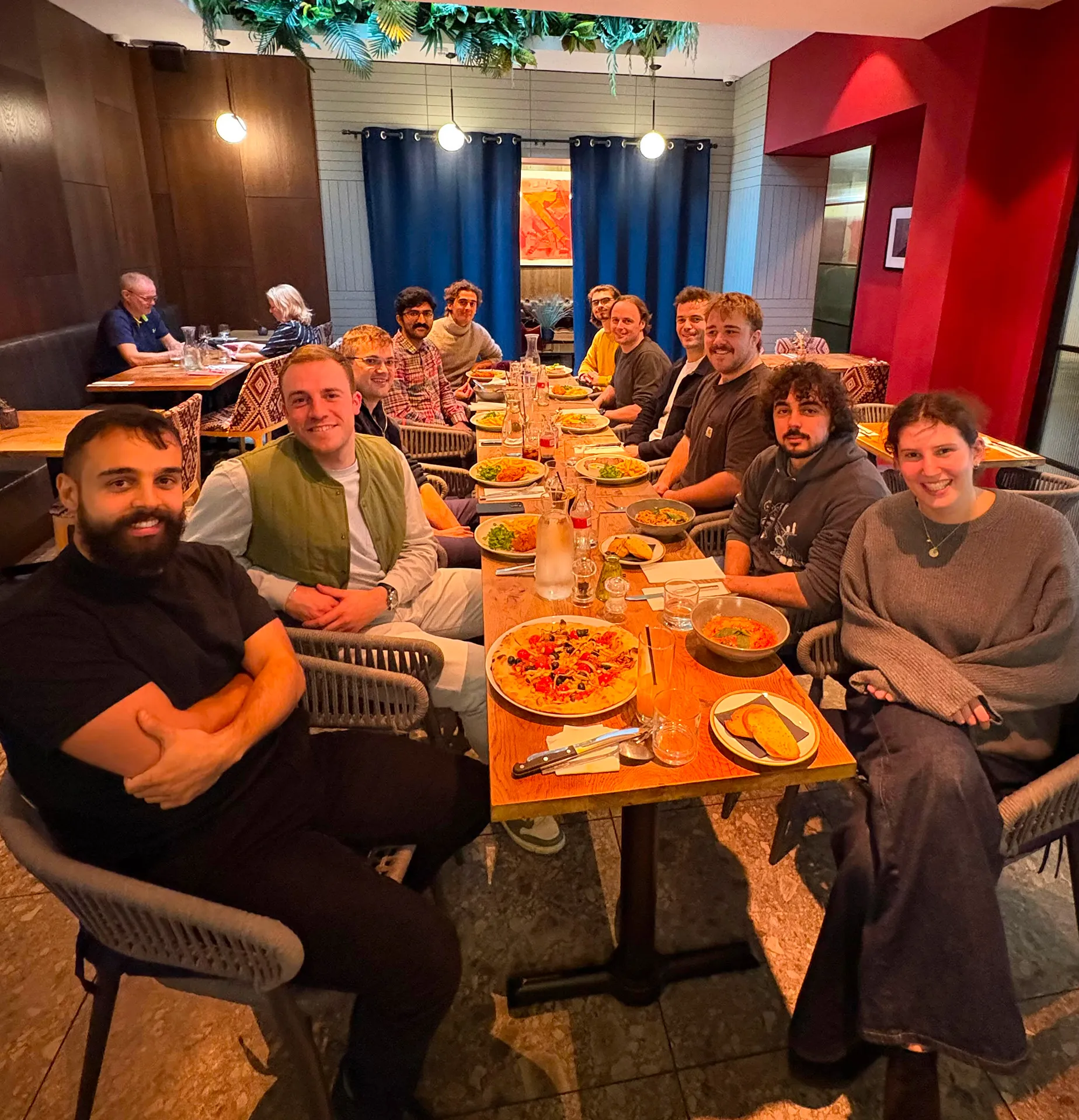 Group of twelve people sitting around a wooden dining table with various dishes including pizza, salads, and drinks in a cozy restaurant.